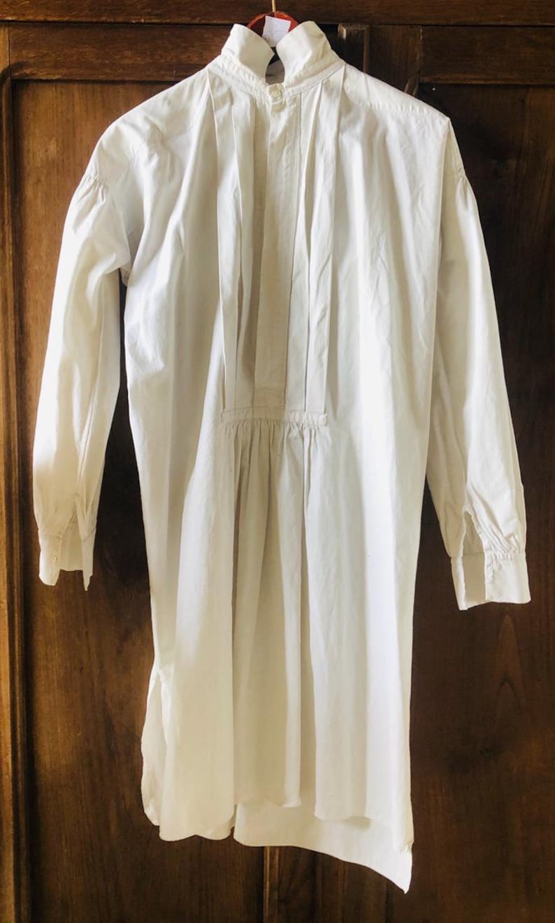 May include: A white cotton shirt with a pleated front and long sleeves. The shirt has a high collar and a button-up closure.