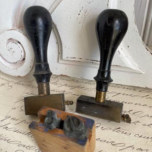 Könnte beinhalten: Zwei antike Stempel mit schwarzen Griffen und Messingstempelköpfen. Ein Stempel ist ein Holzblock mit blauem Rand und zwei Metallstempeln. Die Stempel befinden sich auf einem Vintage-Papierhintergrund.