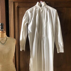 May include: A white cotton shirt with a high collar and long sleeves. The shirt has a gathered waist and a slightly longer back hem.