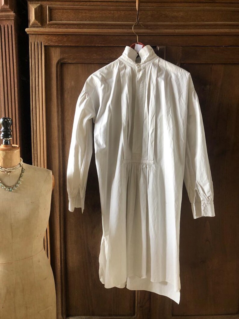 May include: A white cotton shirt with a high collar and long sleeves. The shirt has a gathered waist and a slightly longer back hem.