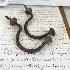 Può includere: Due ganci antichi in metallo curvo con filettature a vite e teste rotonde in ottone invecchiato. I ganci sono marrone scuro con una finitura rustica, su uno sfondo di scrittura vintage.