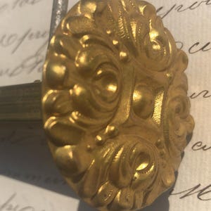 Può includere: Un pomello decorativo in metallo dorato con un design floreale ornato. Il pomello è attaccato a un'asta di metallo.