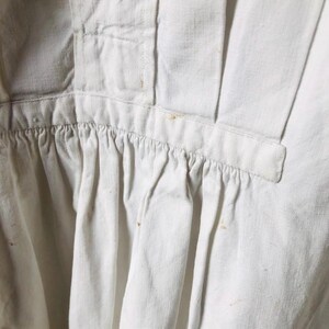 May include: A white cotton garment with a gathered ruffle at the bottom. The garment has a button closure at the neck and a seam down the center back.