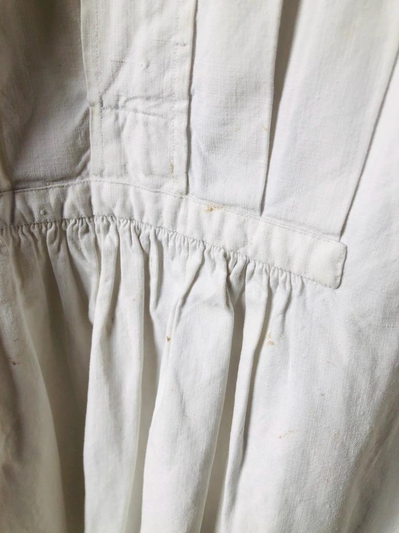 May include: A white cotton garment with a gathered ruffle at the bottom. The garment has a button closure at the neck and a seam down the center back.