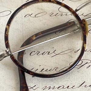 May include: Close-up of vintage round eyeglasses with a tortoiseshell pattern on the frames. The glasses have clear lenses and silver-toned metal arms. The background is a cream-colored paper with handwritten script.