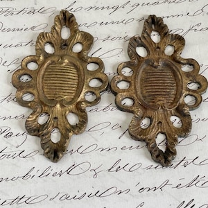 Puede incluir: Dos escudos para muebles de metal en tono latón antiguo a juego, con un diseño calado simétrico y ornamentado, que presenta un medallón ovalado central con estrías horizontales y volutas decorativas en los bordes exteriores.