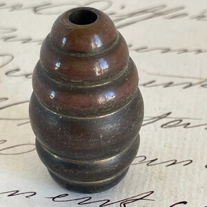 May include: A dark brown, antique-style bead with a layered design. The bead has a cylindrical shape with a hole at the top. The bead's surface has a metallic sheen, suggesting it is made of metal. The bead is likely used for jewelry making or other crafts.