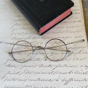 Könnte beinhalten: Antike runde Brille mit dünnen Metallrahmen liegt auf einem handgeschriebenen Dokument. Ein kleines, schwarzes Buch mit goldenen Akzenten und rosa Seitenrändern liegt über der Brille. Das Dokument weist eine elegante Schreibschrift auf.