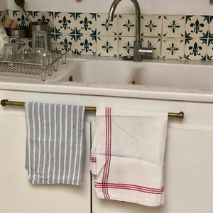 May include: A white kitchen sink with two basins and a chrome tap. Two tea towels hang on a brass bar above the sink. One towel is blue and white striped, the other is white with red stripes.