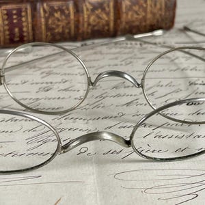 May include: Close-up of antique round eyeglasses with a silver metal frame. The glasses rest on a vintage paper with handwritten script. A stack of old leather-bound books is in the background.