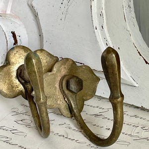 Può includere: Gancio da parete in ottone antico con due ganci curvi. Il gancio presenta una base dal design floreale e una calda patina invecchiata. Lo sfondo include una cornice in legno bianco invecchiato e scritte vintage.