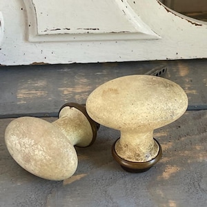 May include: Two antique-style door knobs with an aged, off-white finish. The knobs have a rounded, oval shape and are mounted on a dark metal base. The background features a distressed white painted surface.