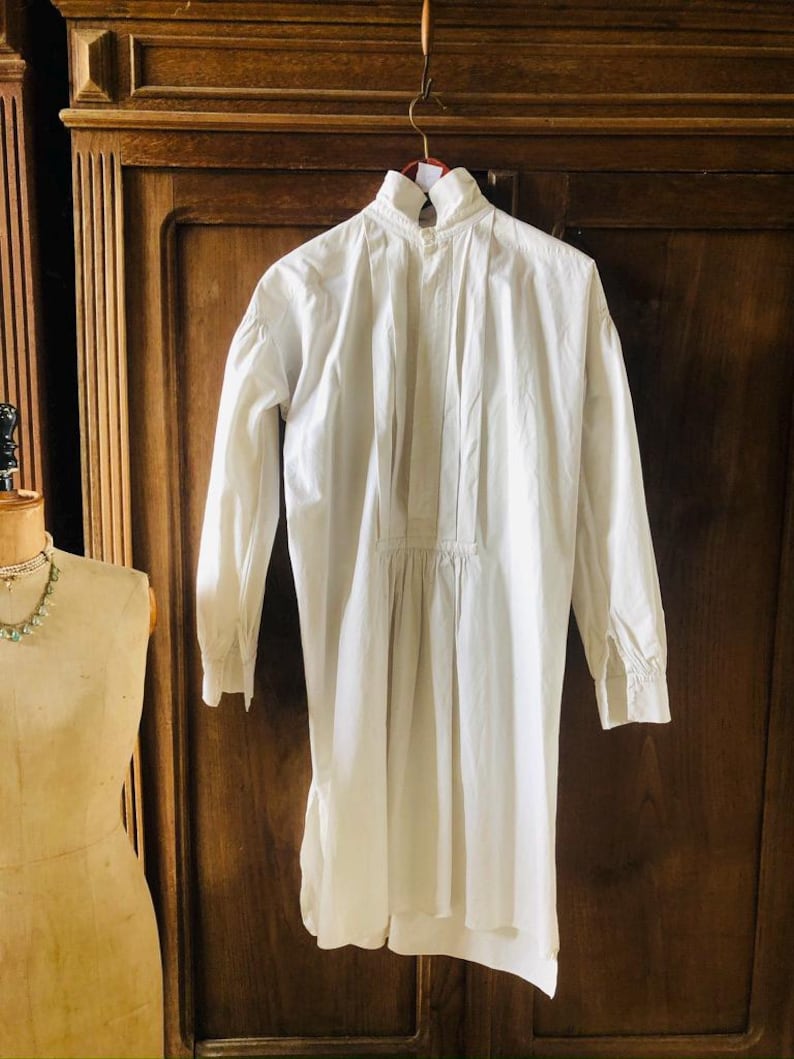 May include: A white cotton shirt with a high collar and long sleeves. The shirt has a gathered waist and a slightly longer back hem.