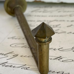 Può includere: Un gancio in ottone vintage con una parte superiore decorativa sfaccettata. Il gancio ha un gambo lungo e sottile e una base circolare. L'ottone ha una calda patina invecchiata, che suggerisce che si tratta di un oggetto antico o vintage. Il gancio è progettato per essere montato su una parete o altra superficie.