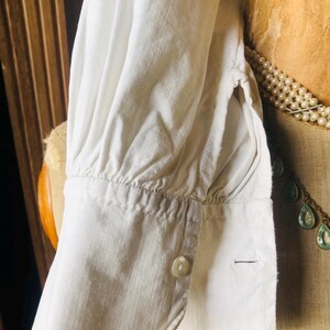 May include: White cotton shirt with a gathered cuff and a single button closure. The shirt is shown on a dress form.