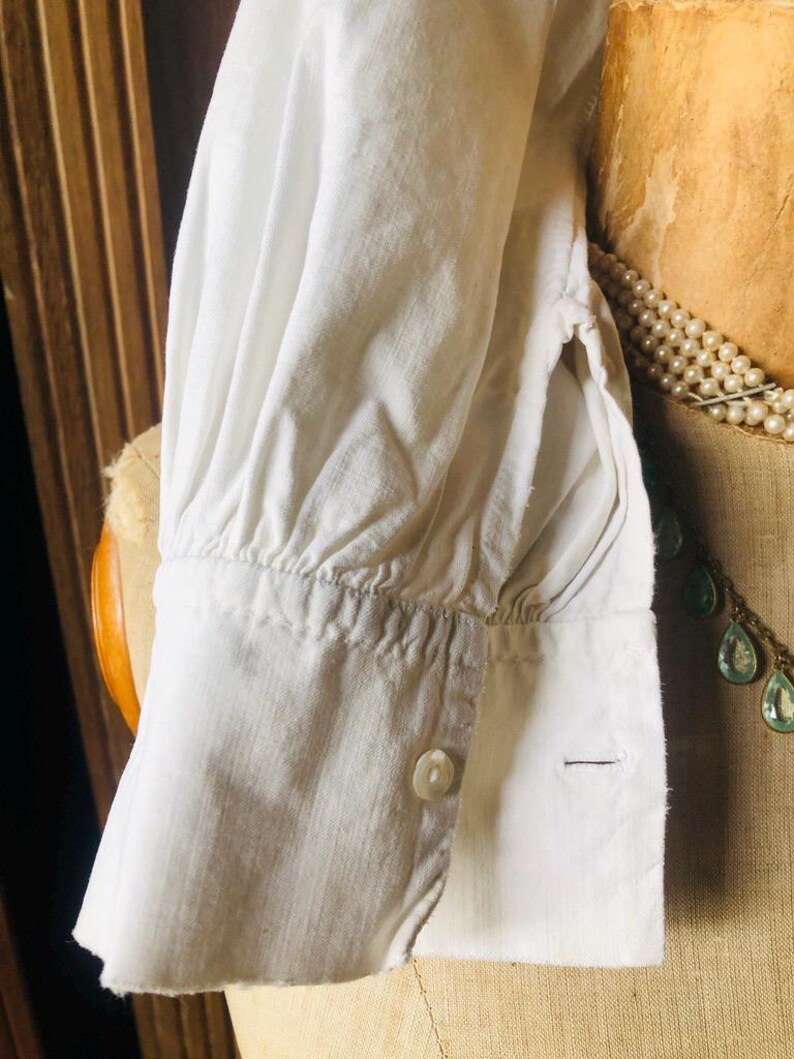 May include: White cotton shirt with a gathered cuff and a single button closure. The shirt is shown on a dress form.