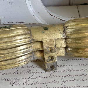 Könnte beinhalten: Ein antikes Möbelbeschlagteil aus goldfarbenem Metall. Das Beschlagteil hat ein dekoratives, symmetrisches Design mit einem zentralen Scharnier und verzierten, geschwungenen Details. Das Teil ist gealtert, mit sichtbaren Gebrauchsspuren.