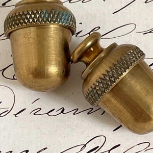 May include: Two brass acorn-shaped objects with a metallic gold finish. Each piece features a smooth, rounded base and a textured, cross-hatched cap with a small knob at the top. The metal shows a slight patina and subtle signs of wear.