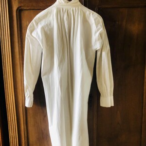 May include: A white long-sleeved shirt with a gathered back detail, hanging on a wooden hanger.