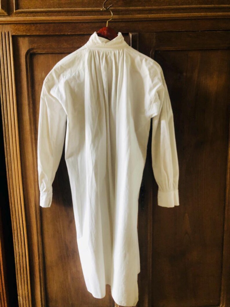 May include: A white long-sleeved shirt with a gathered back detail, hanging on a wooden hanger.