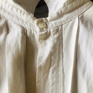 May include: A white linen shirt with a button-up closure and a high collar. The shirt has pleats on the front and is made of a lightweight fabric.