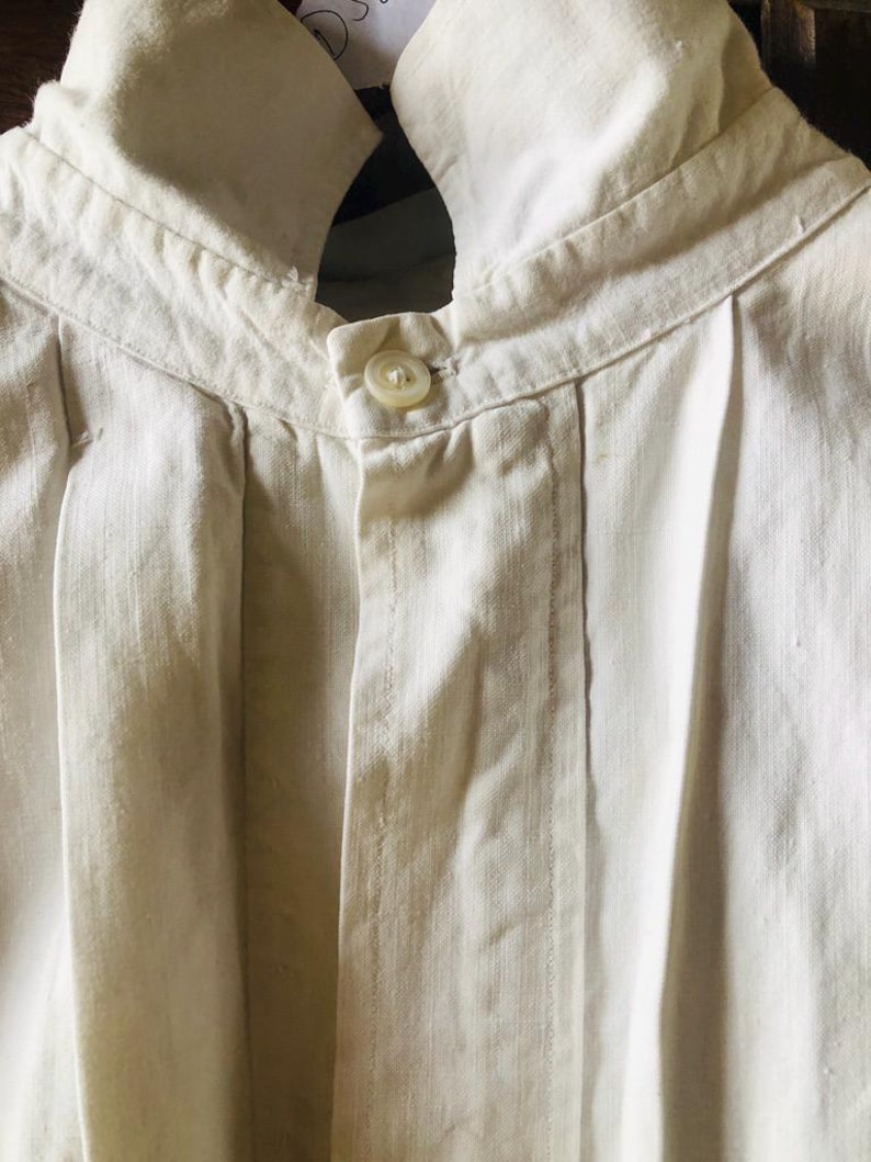 May include: A white linen shirt with a button-up closure and a high collar. The shirt has pleats on the front and is made of a lightweight fabric.