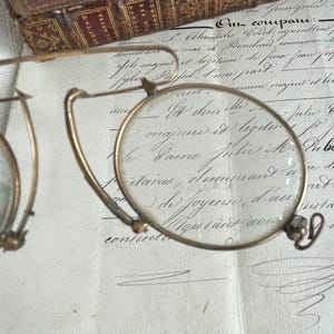May include: Vintage gold-coloured spectacles with circular lenses. The glasses feature a fine frame, set against a backdrop of aged paper with handwritten script and a leather-bound book.