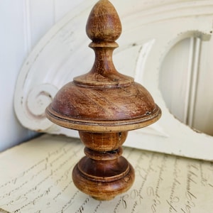 May include: A wooden finial with a rounded top and a fluted base. The finial is a dark brown colour and has a distressed finish.