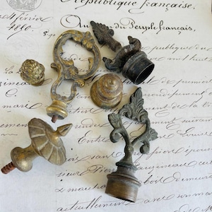 Puede incluir: Una colección de remates de latón antiguos con diseños intrincados. Los remates están dispuestos sobre un trozo de papel con texto escrito a mano en francés.