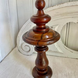 Puede incluir: Objeto decorativo de madera torneada con acabado marrón oscuro. El objeto tiene una base redonda, un pedestal, una sección media ancha, plana y circular, y una parte superior más pequeña y escalonada con un remate redondeado. Fondo blanco.