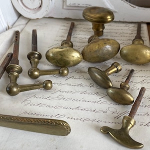 Puede incluir: Una colección de herrajes antiguos de latón, que incluye pomos y manijas de puertas, dispuestos sobre papel vintage con escritura manuscrita. Las piezas de ferretería varían en forma y tamaño, mostrando un aspecto envejecido.