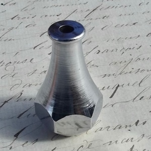 Może przedstawiać: Srebrny metalowy korek do butelki z sześciokątną podstawą i gładkim, zaokrąglonym wierzchołkiem. Korek ma mały otwór w środku wierzchołka.