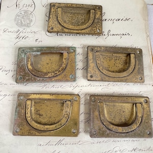 Puede incluir: Seis tiradores de cajón de latón antiguo con forma rectangular redondeada y un recorte de asa semicircular. Los tiradores tienen una pátina desgastada y envejecida.