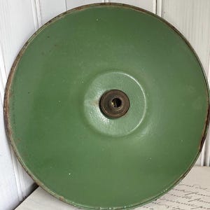 May include: A round, sage green enameled metal lamp shade with a smooth, slightly glossy finish. The shade features a central circular opening with a dark, metallic socket fitting. The outer rim shows signs of wear with visible rust along the edge.