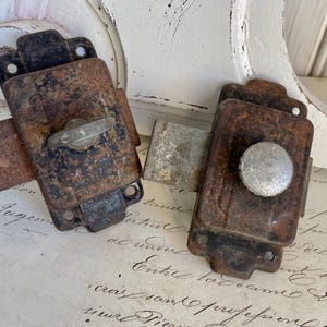 May include: Two antique, rusty metal latches with decorative details. One latch has a small, round knob, and the other has a flat, lever-style handle. Both have mounting holes for screws. The latches are set against a backdrop of aged paper.