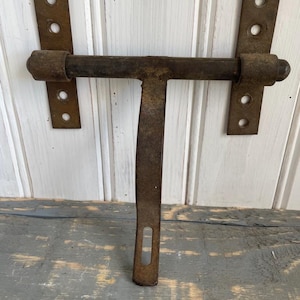 May include: A vintage metal door latch with a rusty finish. The latch features a T-shaped design, with a horizontal bar and a vertical handle. Two rectangular plates with holes are on either side of the bar. The metal has a weathered look.