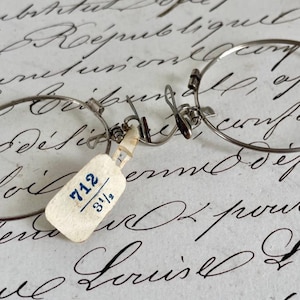 May include: A pair of silver-toned metal pince-nez spectacles featuring thin circular frames and a central bridge mechanism. A small, off-white rectangular paper tag is attached, displaying the blue handwritten text 712 3 1/2.