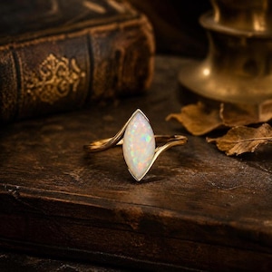 May include: A gold ring featuring a marquise-cut opal gemstone with iridescent flashes of color. The band has a bypass design that curves around the central stone, which is set vertically in a simple metal frame.