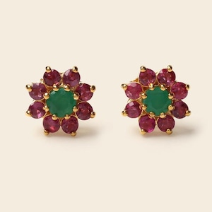 May include: A pair of gold stud earrings featuring a central round green gemstone surrounded by a halo of eight small round red rubies. The floral-shaped design is set in yellow gold, creating a vibrant contrast between the green and red stones.