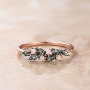 Op de afbeelding: Een roségouden ring met een cluster van donkergroene, marquise-geslepen stenen en kleine ronde witte opalen, gerangschikt in een bladvormig patroon bovenop de dunne, gepolijste metalen ringband.