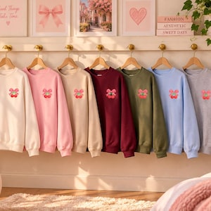 May include: A collection of eight crewneck sweatshirts in white, light pink, beige, maroon, olive green, light blue, gray, and black. Each sweatshirt features a small embroidered graphic on the left chest of two red cherries tied with a pink bow.