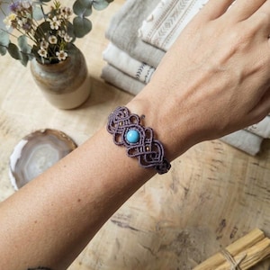 May include: A macrame bracelet featuring a central round turquoise stone set within intricate dark grey knotted cord patterns, accented with small metallic beads. The design showcases a symmetrical woven structure that wraps around the wrist.