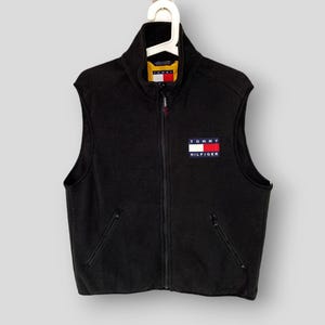 May include: A black fleece vest featuring a full-length front zipper, a high collar, and two zippered side pockets. The Tommy Hilfiger logo is embroidered on the left chest in white, red, and navy blue, with a yellow inner collar lining.