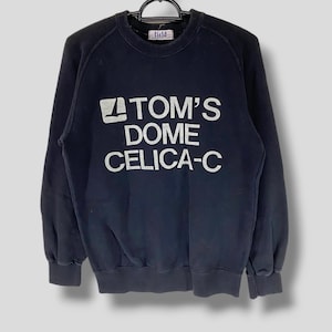 May include: A black long-sleeve crewneck sweatshirt featuring a white graphic print on the chest that reads TOM'S DOME CELICA-C with a small logo icon to the left. The garment has ribbed cuffs and a ribbed hem, hanging on a black hanger.