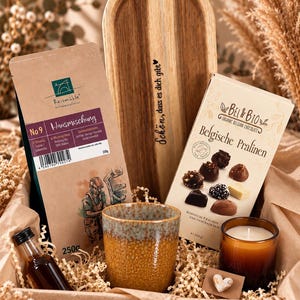 Op de afbeelding: Een geschenkset met een zak van 250g Reis Mühle koffie, een houten dienblad met de tekst "Schön, dass es dich gibt", een doos Bel & Bio Belgische chocolaatjes, een gespikkelde bruine keramische mok, een klein glazen flesje en een geurkaars.