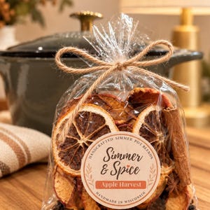 May include: A clear plastic bag tied with a jute twine bow containing dried orange slices, apple slices, cinnamon sticks, and star anise. A circular label reads Handcrafted Simmer Pot Blends, Simmer & Spice, Apple Harvest, Handmade in Wisconsin.