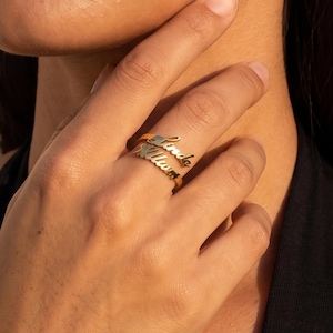 May include: A gold-toned ring featuring two stacked bands, each displaying a custom name in a cursive script font. The metal has a polished, reflective finish, and the design consists of two distinct, connected nameplates worn on a single finger.