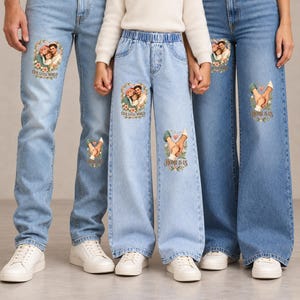 May include: Three pairs of light blue denim jeans featuring custom printed graphics. The designs include a family portrait illustration with the text "OUR LITTLE WORLD" and a close-up of hands joined together with the text "HOME IS US".