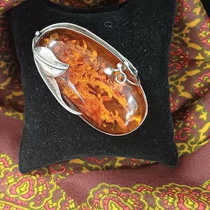 May include: An oval-shaped brooch featuring a large, polished amber stone with visible internal inclusions. The amber is encased in a silver setting that includes a stylized leaf motif on one side and a small, coiled wire detail on the other.