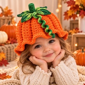 May include: A bright orange crocheted hat shaped like a pumpkin, featuring a textured ribbed design, a green stem at the top, and decorative green crocheted leaves and a curly vine extending from the center of the crown with a ruffled brim.